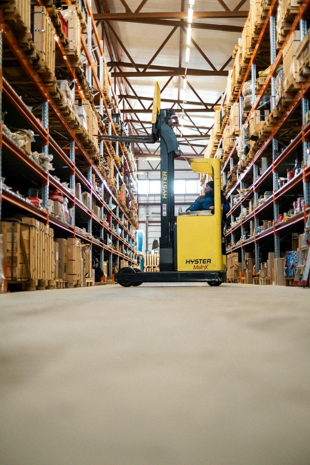 category-02 Warehouse Worker Organizing Inventory on a Ladder A warehouse worker in blue overalls and a beanie is standing on a metal ladder, reaching for a package on a high shelf. The warehouse is filled with industrial shelves stocked with boxes and various supplies. The image captures the essence of logistics, inventory management, and manual labor in a structured warehouse environment.